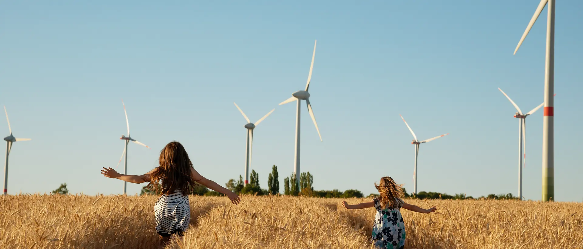 Duas meninas a correr num milharal com torres eólicas ao fundo