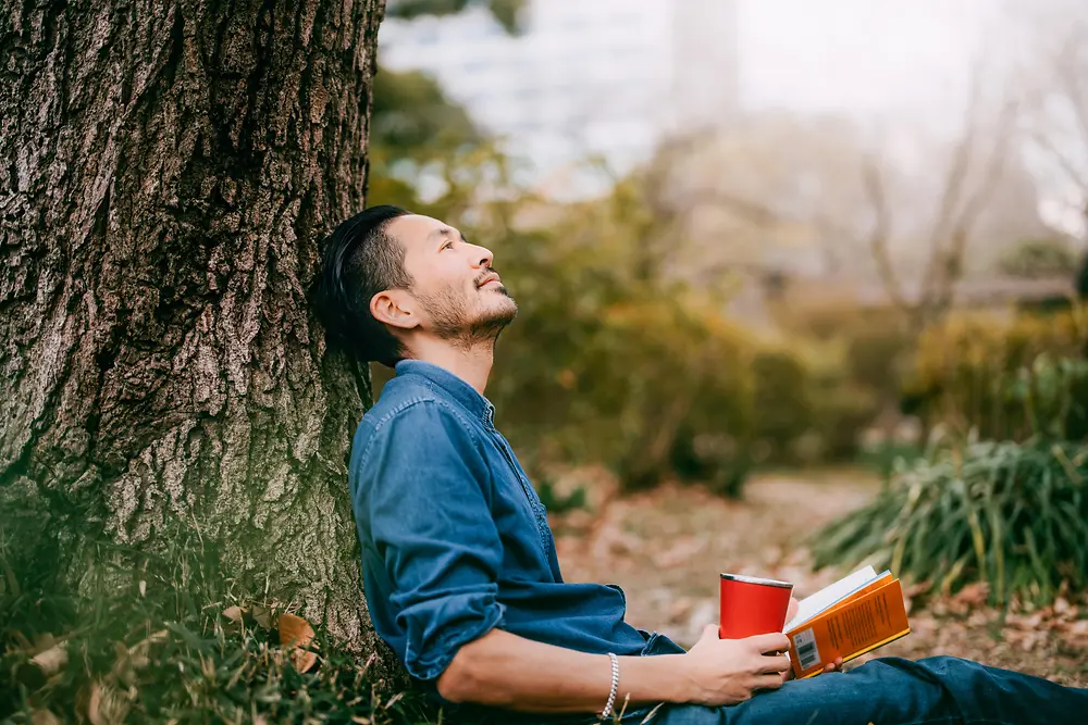 Um homem com um livro encostado a uma árvore